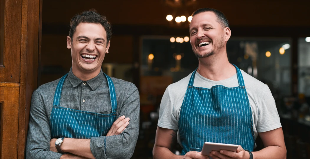 Two smiling cafe owners in aprons standing at their shop entrance